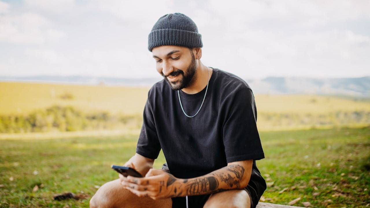 De moderne man in casual stijl met tattoos, zwarte outfit en muts, zittend buiten met smartphone in de hand.