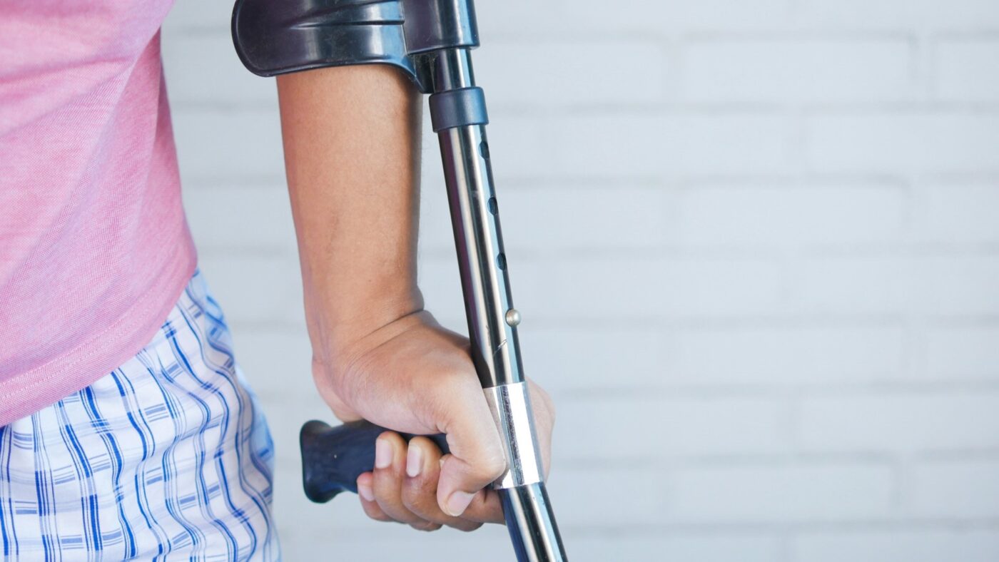 Persoon met blessure ondersteunt zichzelf met een kruk tegen een lichte achtergrond