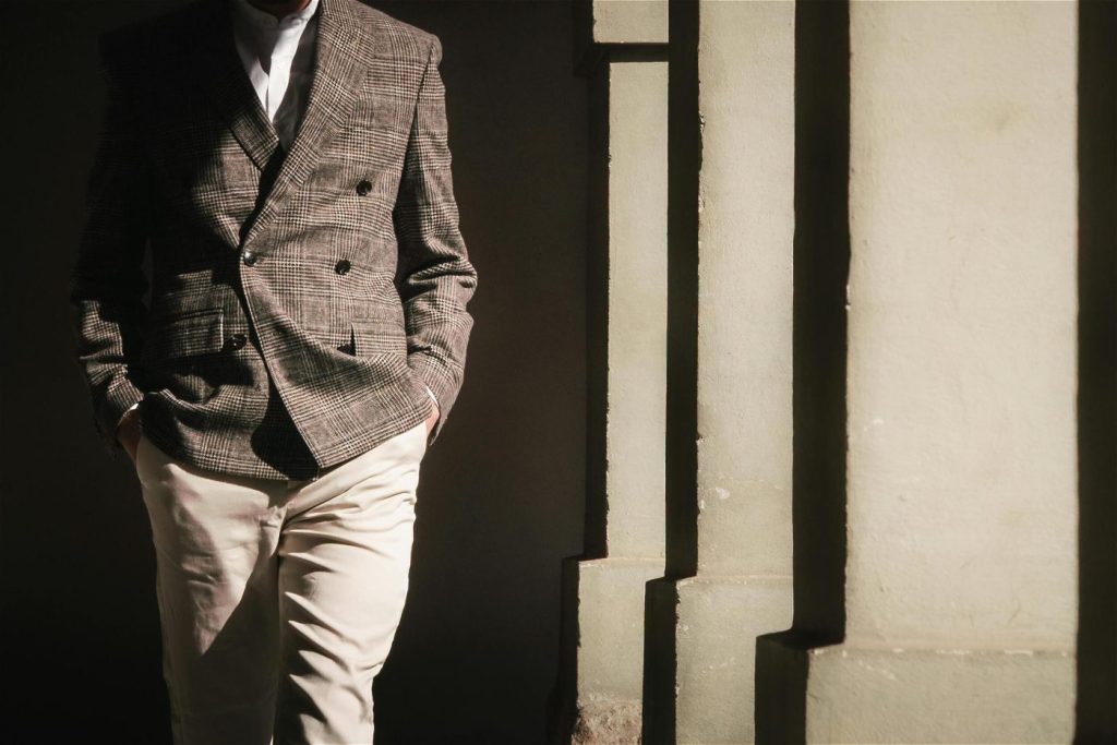 Man in colbert en lichte broek staat bij muur met handen in zakken