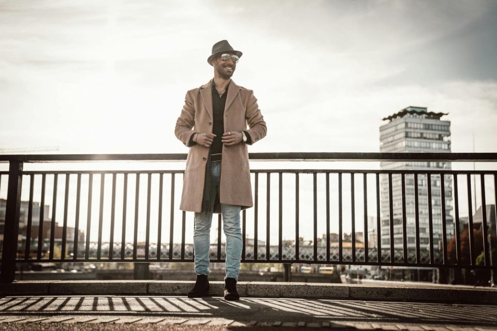 Man met hoed en lange jas staat op brug met stadsgebouw op de achtergrond