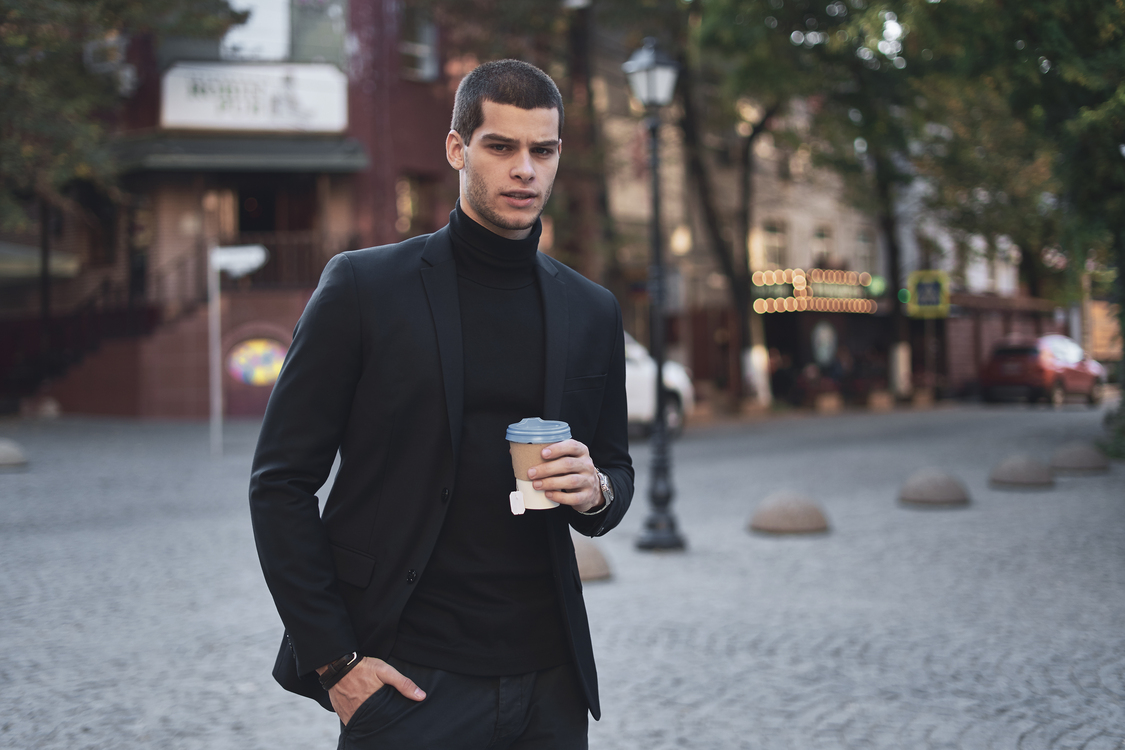 Jonge man in zwarte coltrui en blazer met koffie in een stedelijke omgeving.