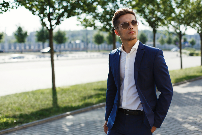Jonge man in blauw pak met zonnebril loopt door een park.